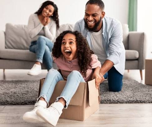 A family moving into their rental, the daughter laughing as her dad pushes her across the floor in a moving box